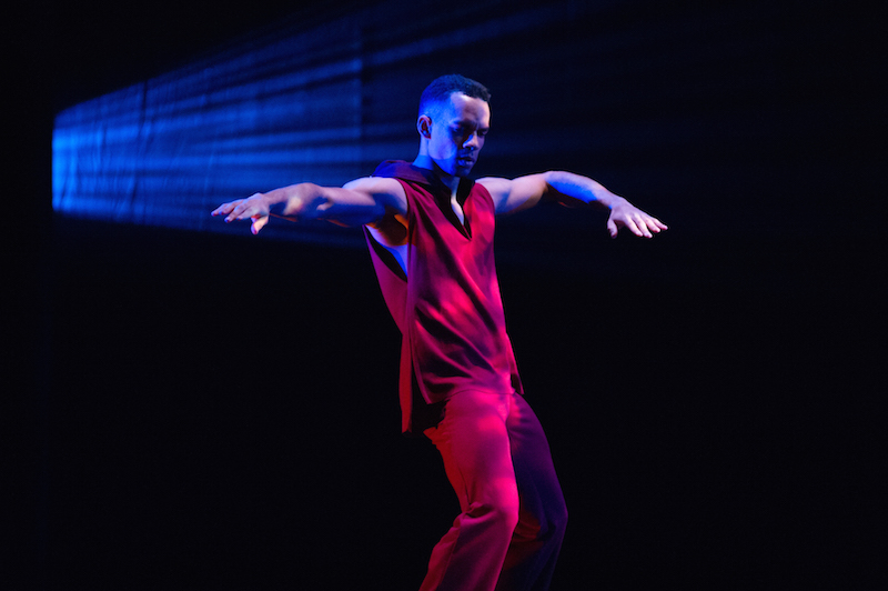 A dancer in a red tunic and pants bends his knees and extends one arm while his other is bent at an angle.